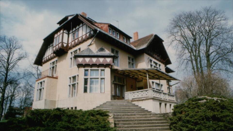 A large, multi-story house with a steep roof, a balcony, and a set of stairs leading to the entrance.