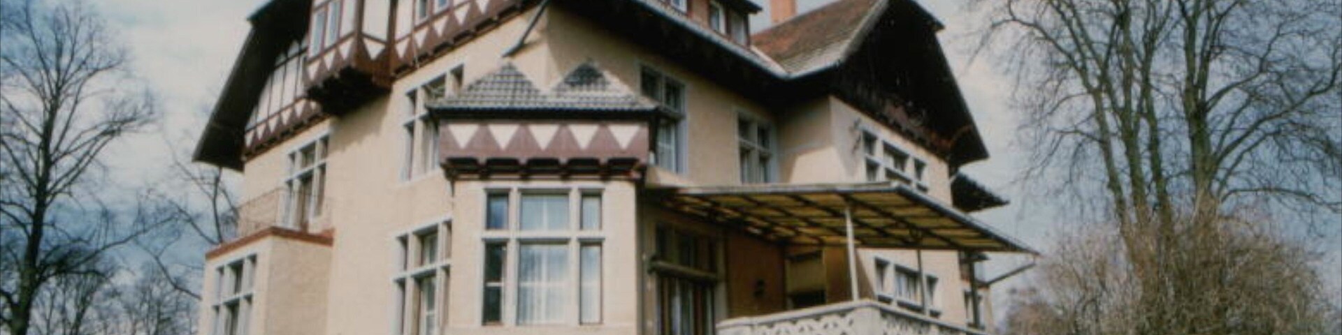 A large, multi-story house with a steep roof, a balcony, and a set of stairs leading to the entrance.