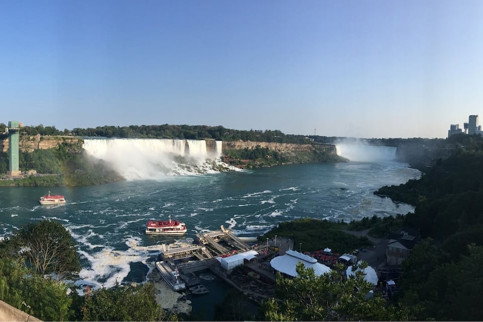 A river with a waterfall, boats, a bridge, and a city skyline with a tower.