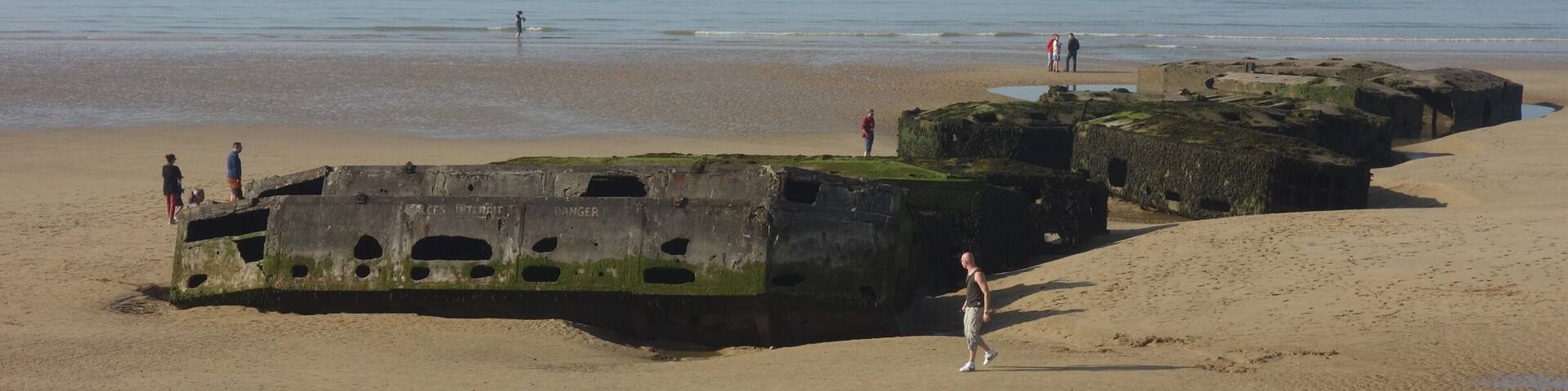 A beach with large concrete structures covered in seaweed, people walking, and distant boats on the water.