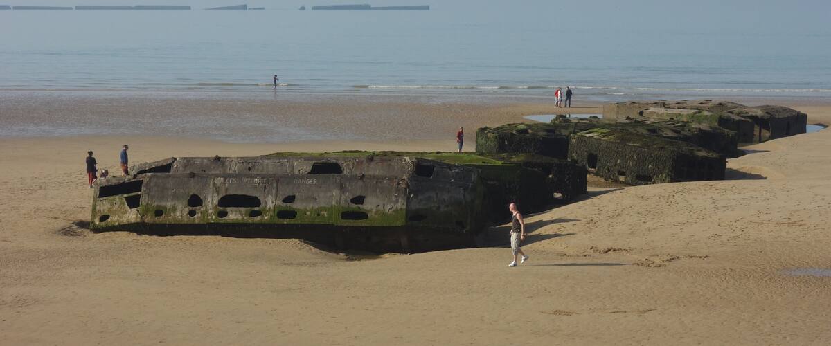 A beach with large concrete structures covered in seaweed, people walking, and distant boats on the water.