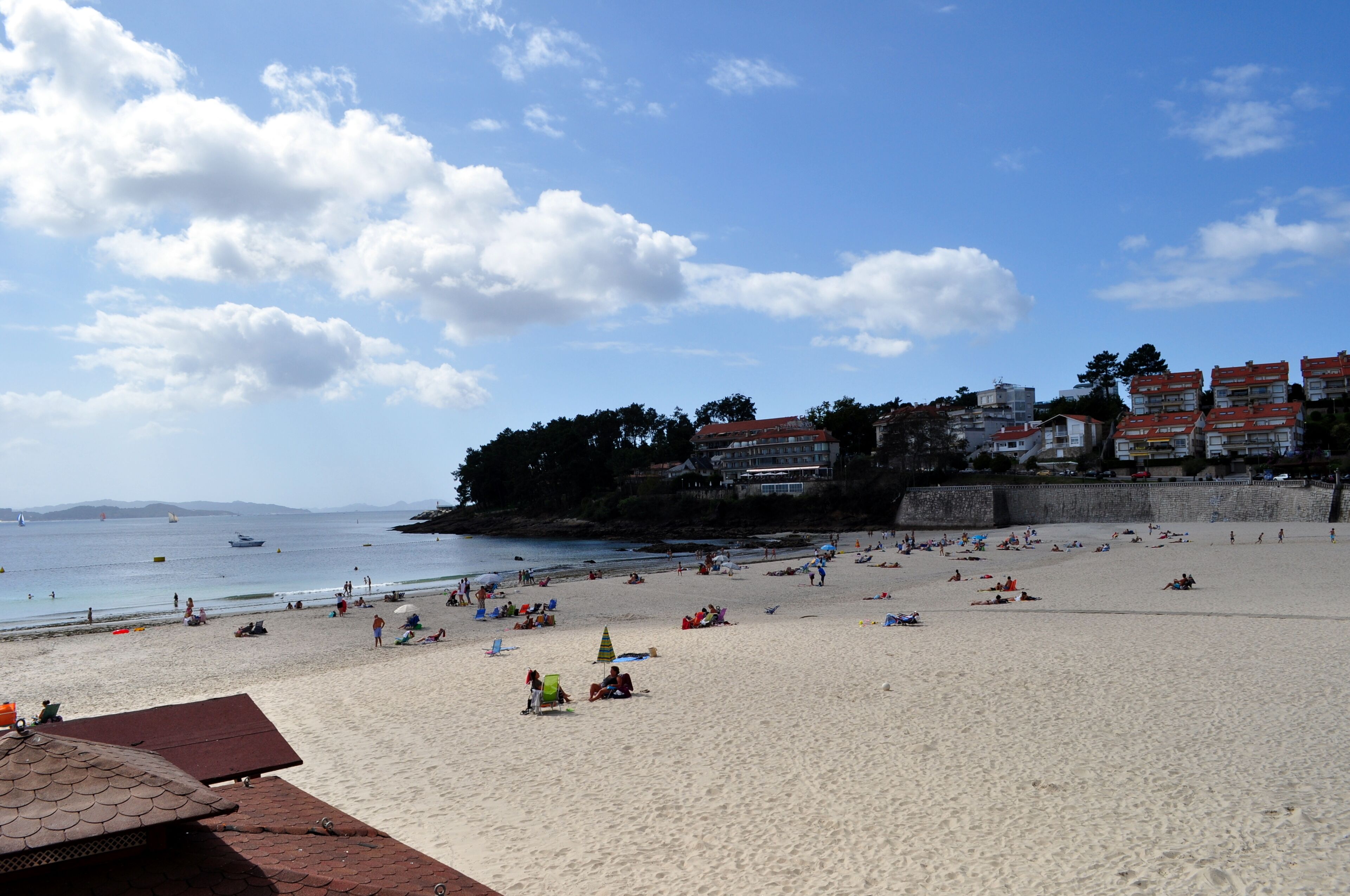 A beach with people sunbathing and swimming, a clear sky, and buildings in the background.