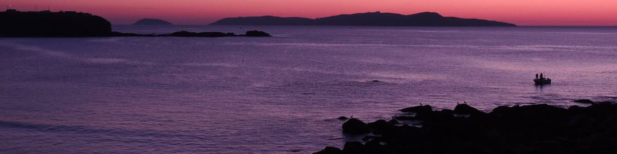 Ein Strand bei Sonnenuntergang mit ruhigem Wasser, Felsen und in der Ferne Inseln.