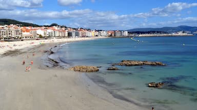 Una playa con agua cristalina, afloramientos rocosos y edificios al fondo.