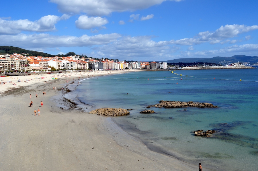 Una playa con agua cristalina, afloramientos rocosos y edificios al fondo.