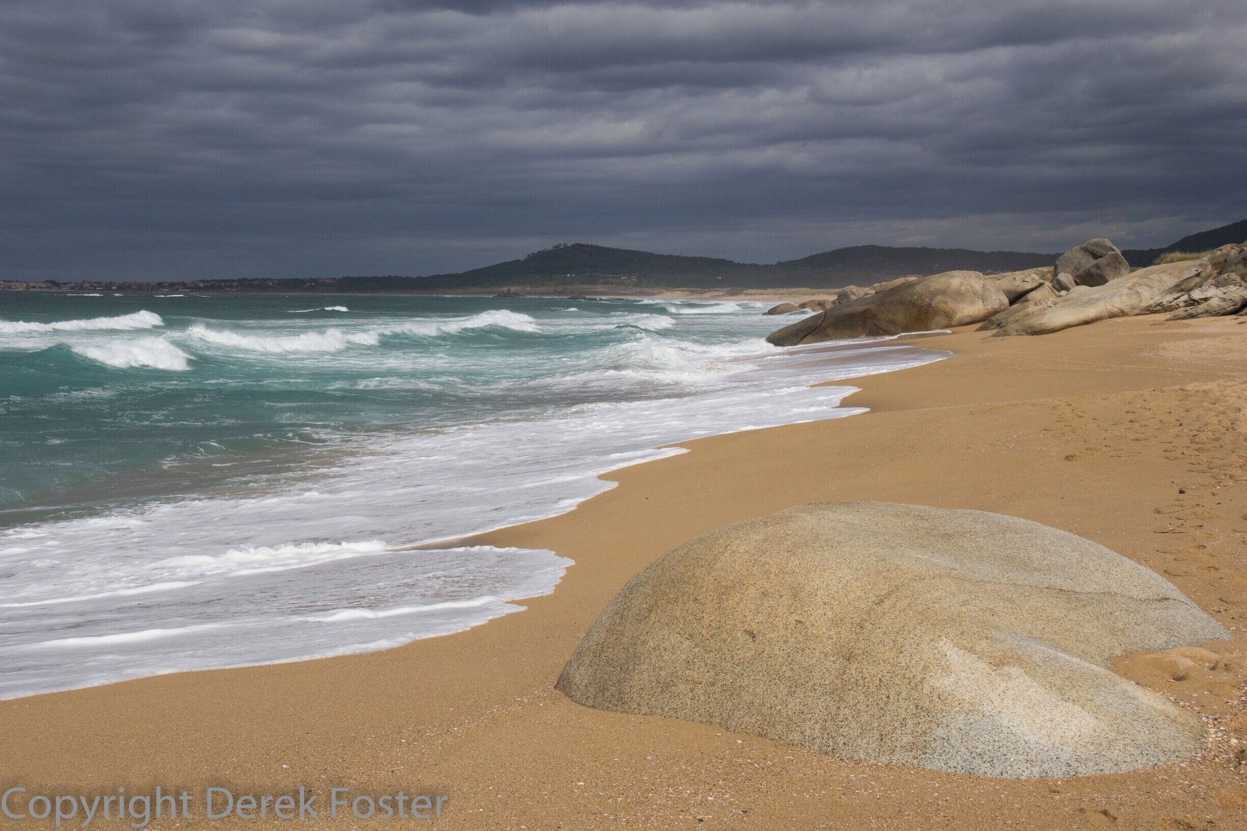 A beach with waves, a large rock, and a cloudy sky.