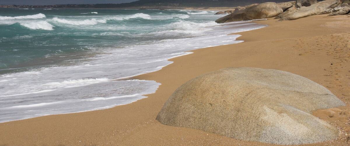 A beach with waves, a large rock, and a cloudy sky.