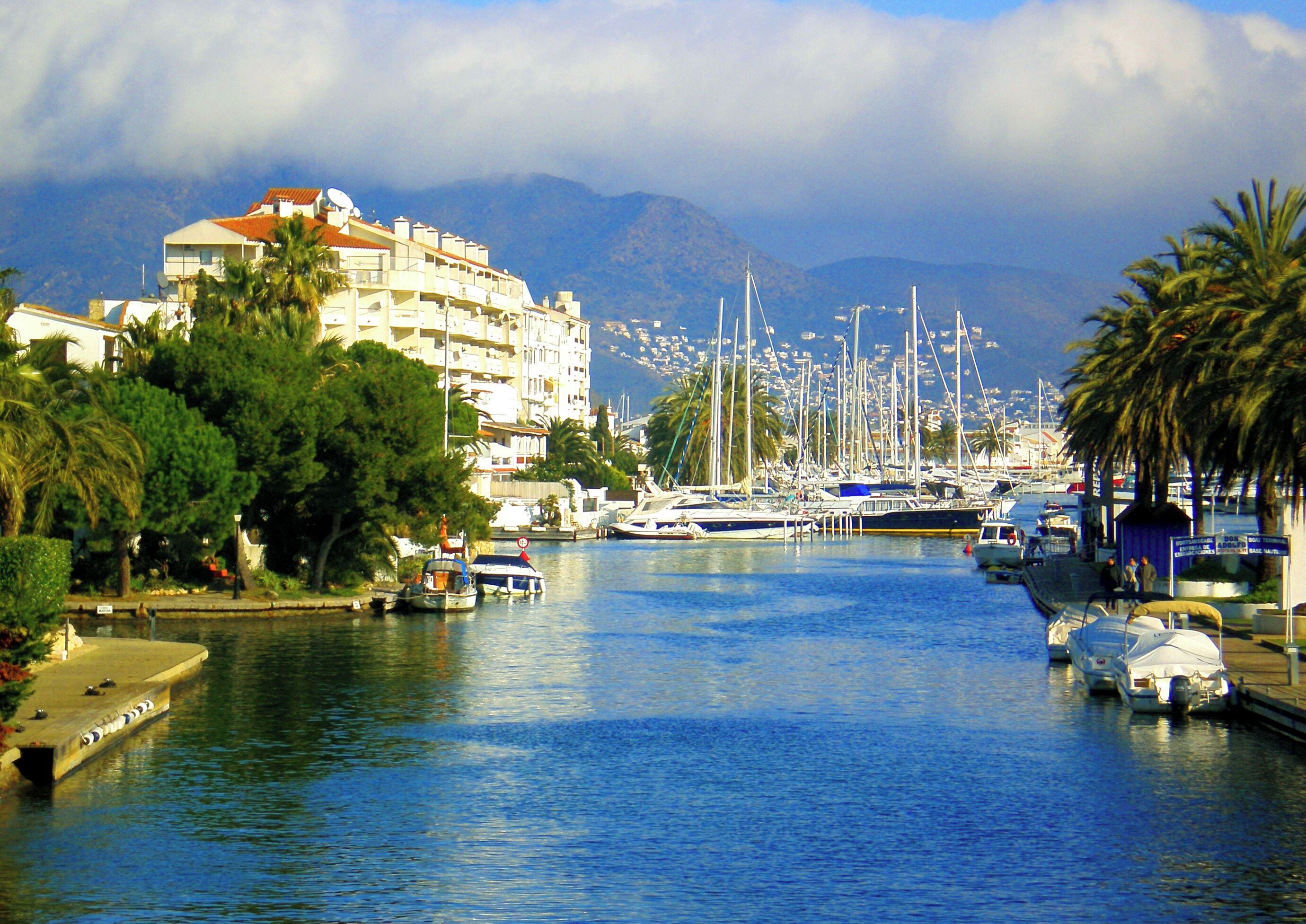 A marina with boats docked, a building with a red roof, and mountains in the background.