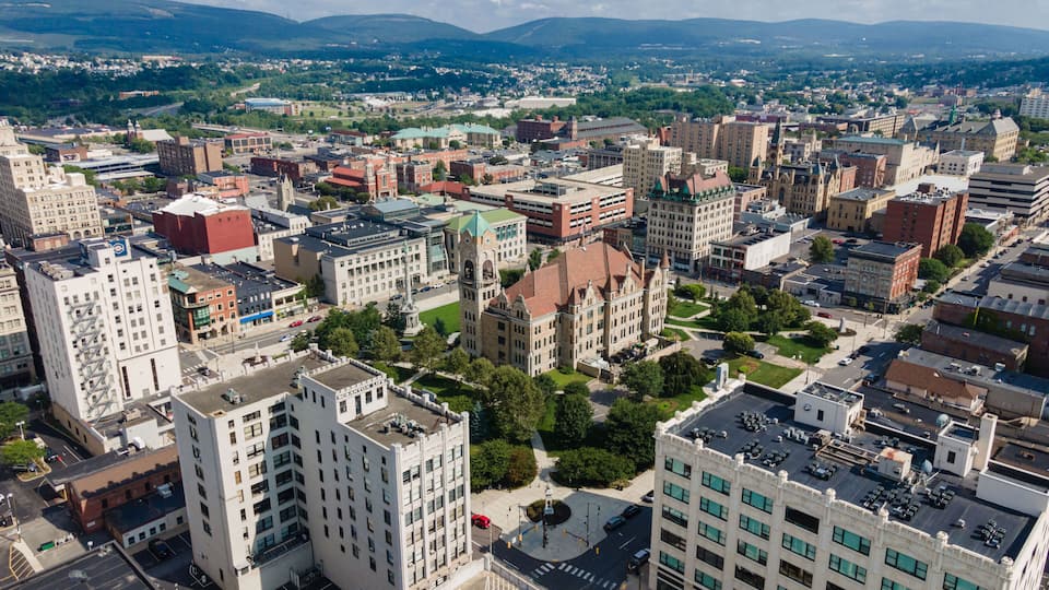An aerial view of a city with historic architecture, green spaces, and a mountainous backdrop.