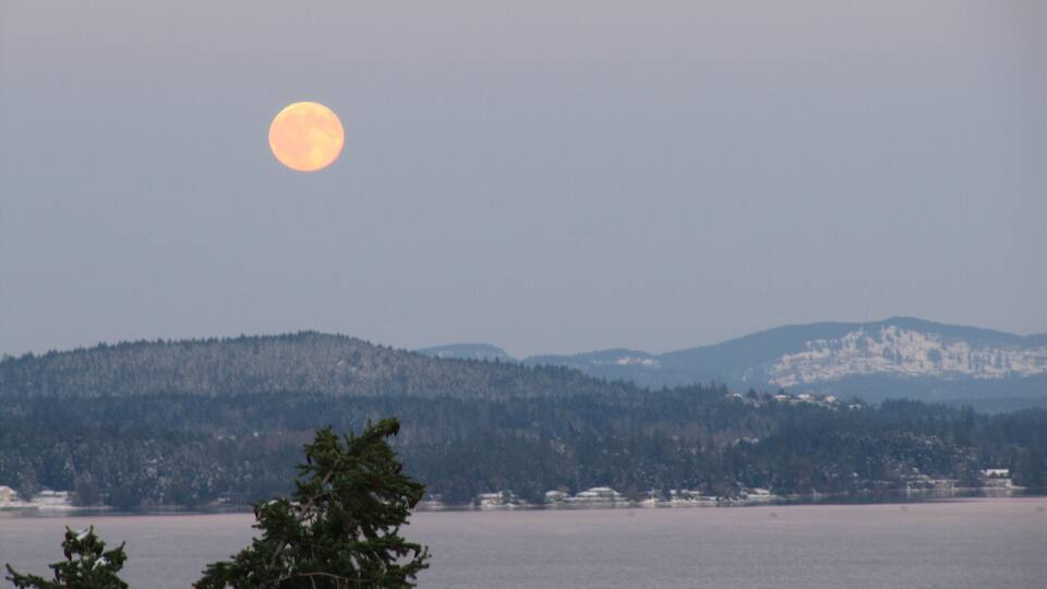 A large moon over a calm lake with mountains in the background.