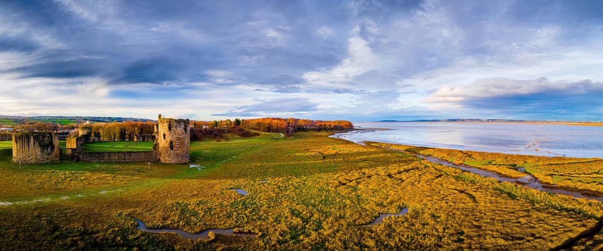 A castle ruins by a river with autumn foliage and a cloudy sky.