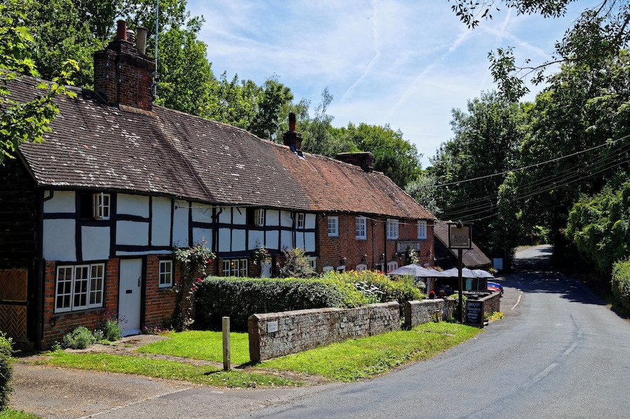 A traditional half-timbered house with a brick chimney, a stone wall, and a signboard.