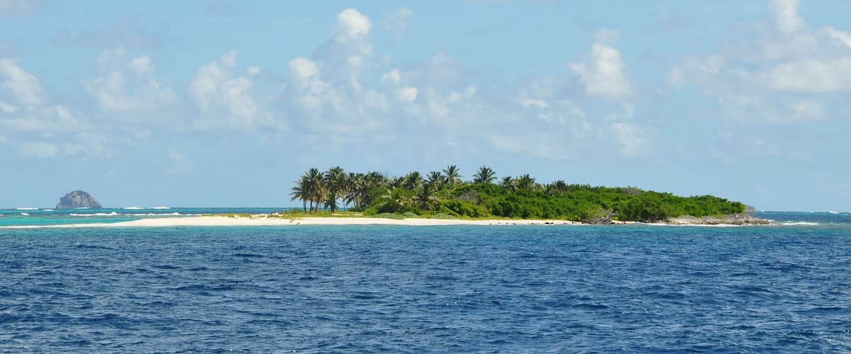 A sailboat on turquoise waters, a sandy beach, and a small island with palm trees.