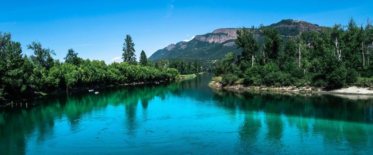 A river with clear turquoise water, surrounded by dense green forest and a mountain in the background.