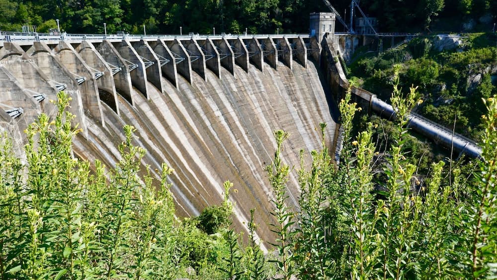 A large dam with multiple spillways, surrounded by greenery and rocky terrain.