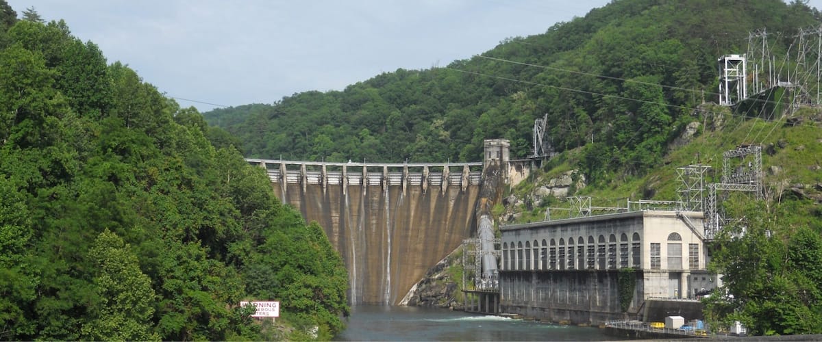 A large dam with multiple gates, surrounded by lush green forest and hills.