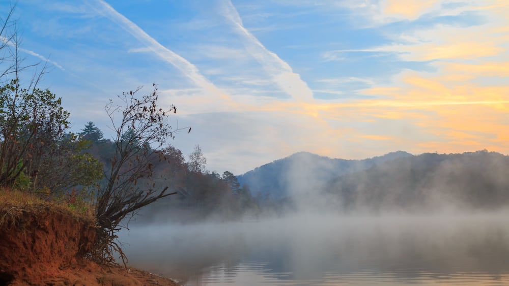 Ein ruhiger See mit einem nebligen Vordergrund, einem klaren Himmel mit Kondensstreifen und einem bewaldeten Gebirgszug im Hintergrund.