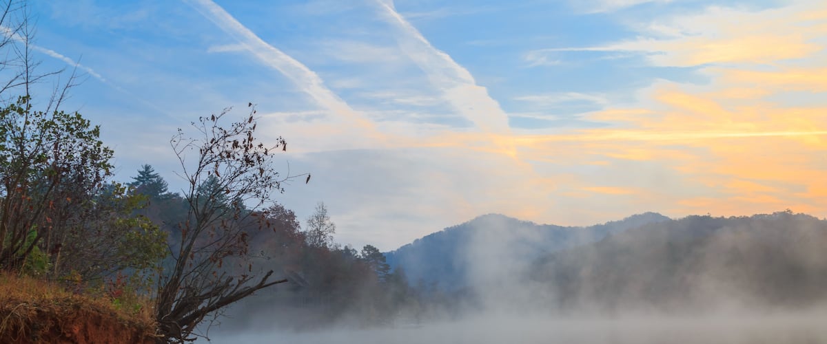 Un lac paisible, avec un premier plan enveloppé de brume, un ciel dégagé traversé par des traînées de condensation et, au loin, une chaîne de montagnes boisée.
