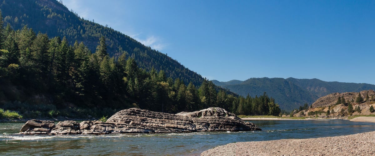A river with a large rock formation in the middle, surrounded by dense forests and mountains.