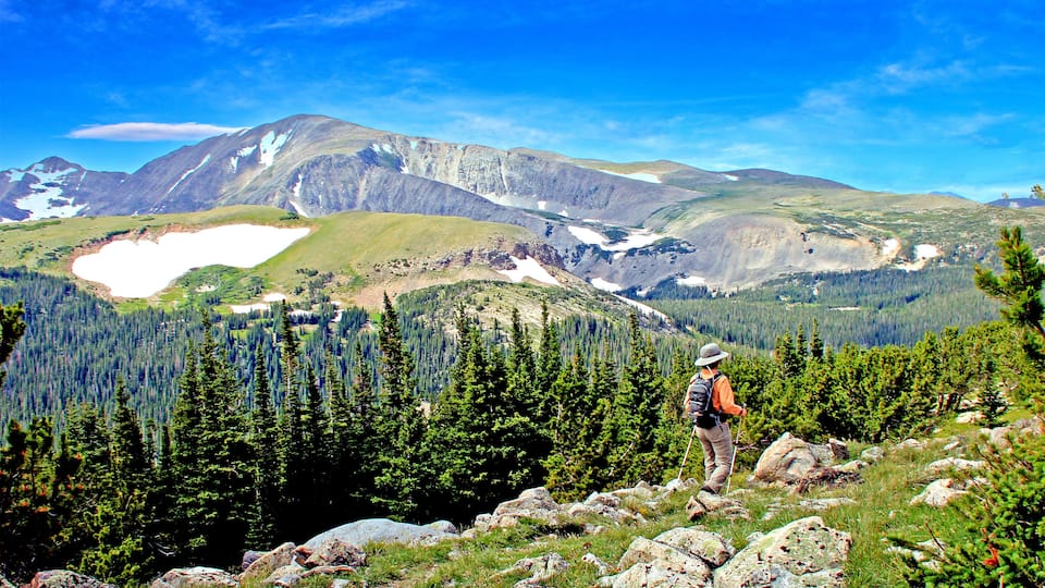 A hiker on a mountain trail with a lake and forested mountains in the background.