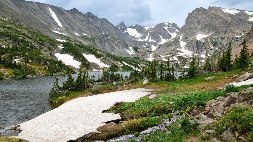 A mountain landscape with a lake, snow patches, and green vegetation.