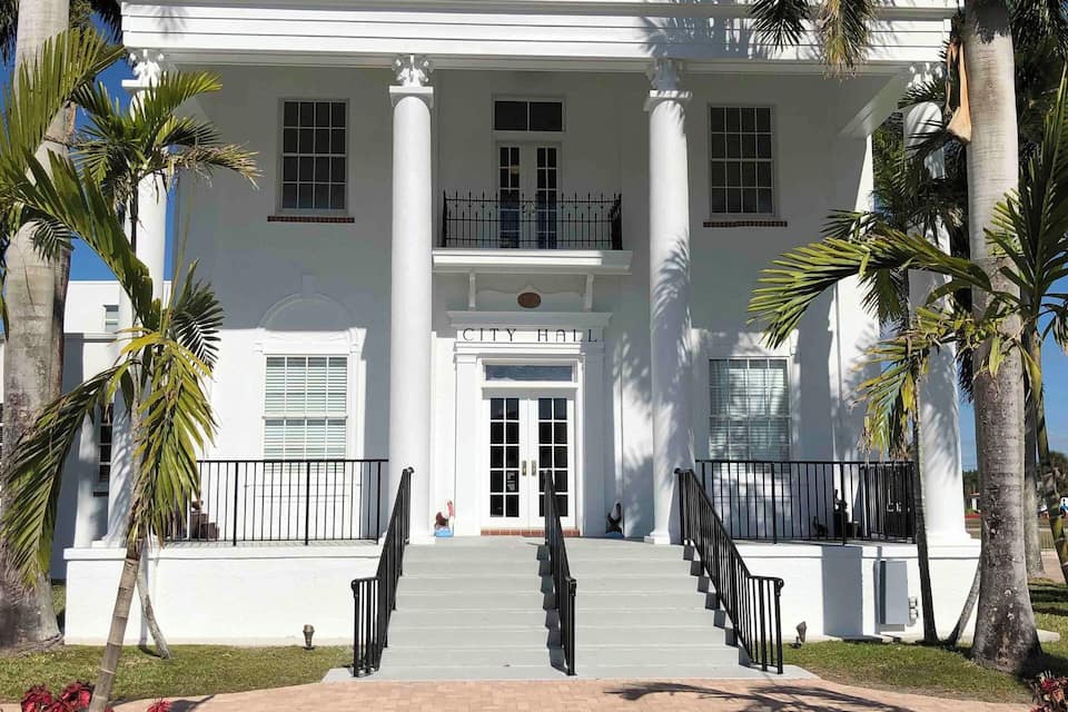 A white building with columns and a balcony, labeled "City Hall," surrounded by palm trees.