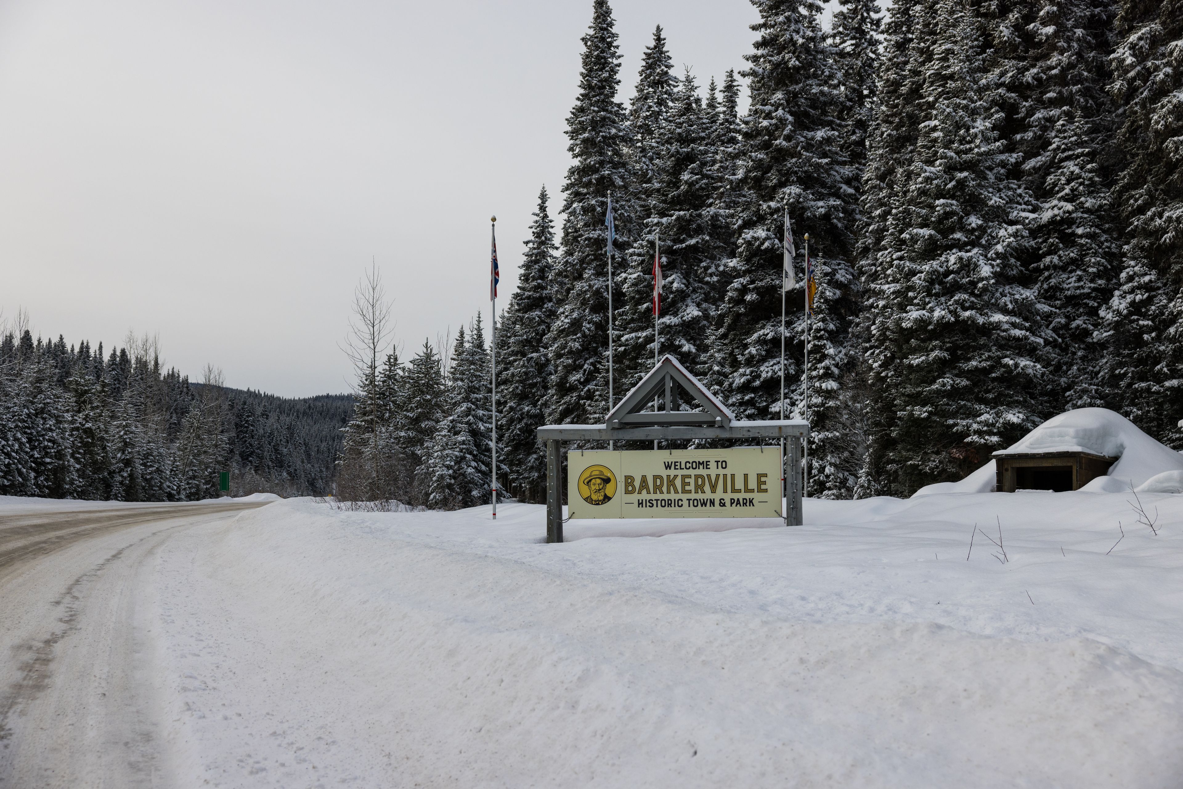 A snow-covered road leading to a sign welcoming visitors to Barkerville, a historic town and park.