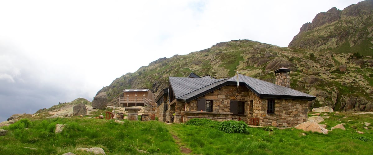 A stone house with a sloped roof surrounded by lush green grass and rocky mountains in the background.