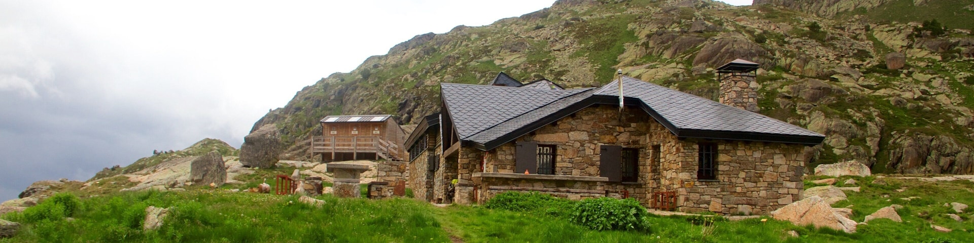 A stone house with a sloped roof surrounded by lush green grass and rocky mountains in the background.