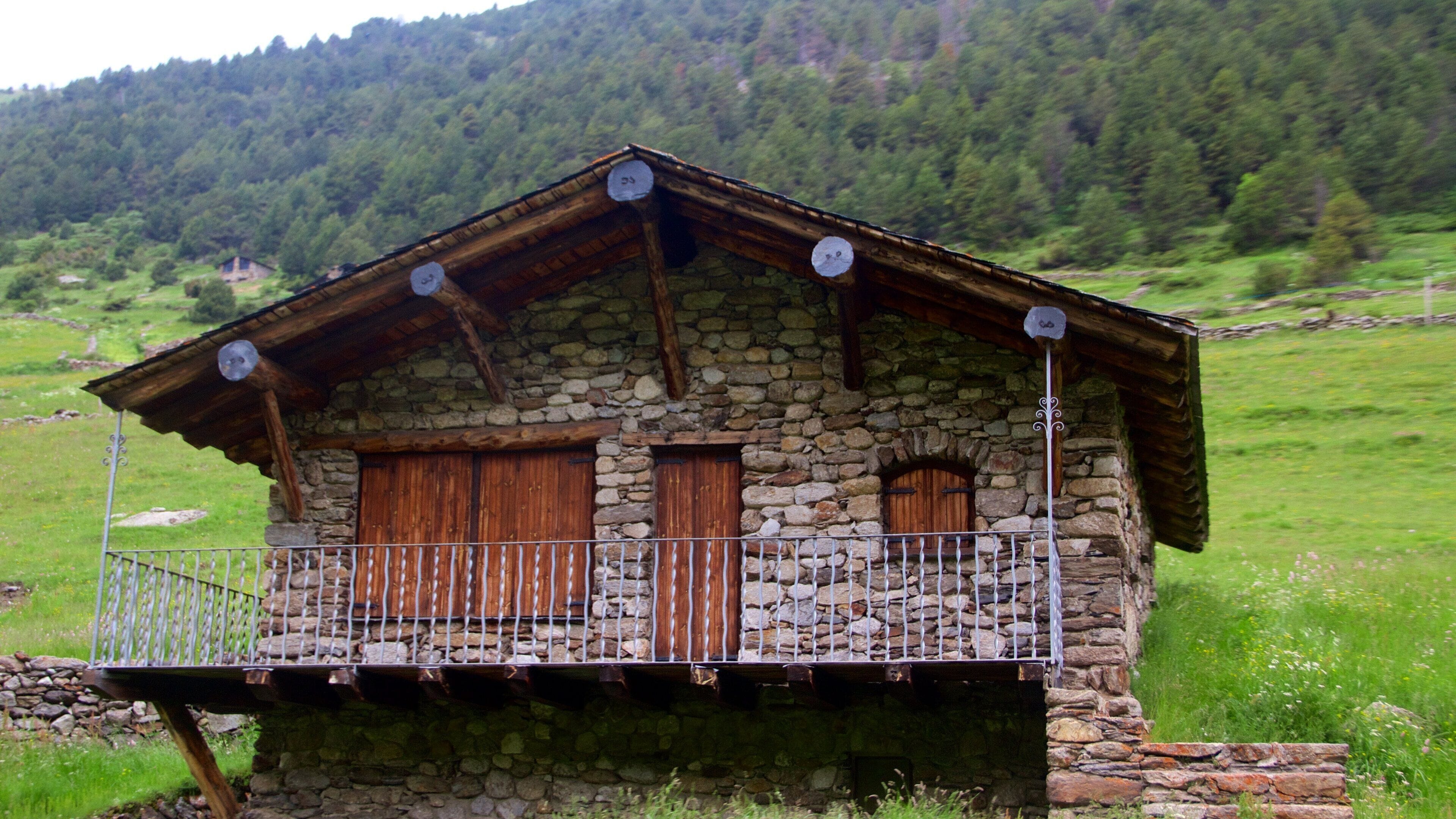 A stone house with wooden doors and windows, a balcony with a metal railing, and a mountainous backdrop.
