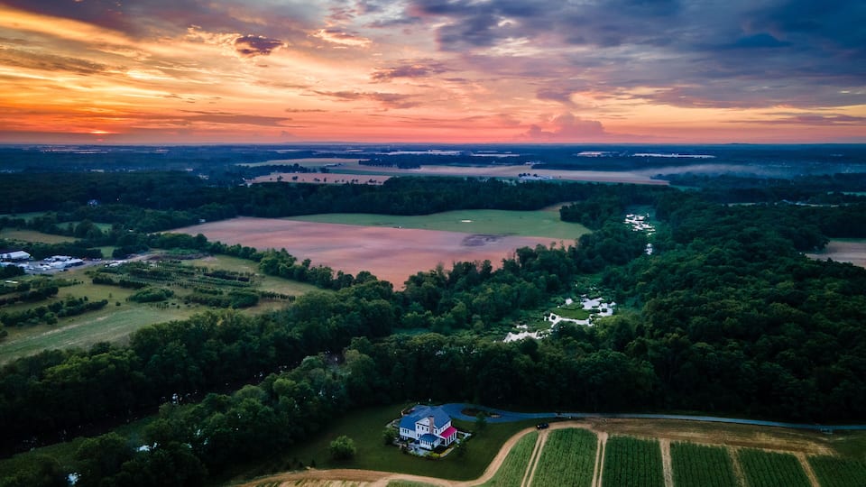 An aerial view of a rural landscape with fields, a large body of water, and a house surrounded by trees.