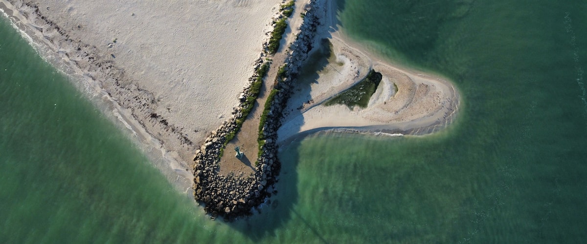 An aerial view of a coastal area with a long, narrow strip of land, a small island with a curved shoreline, and a clear demarcation between the land and the water.