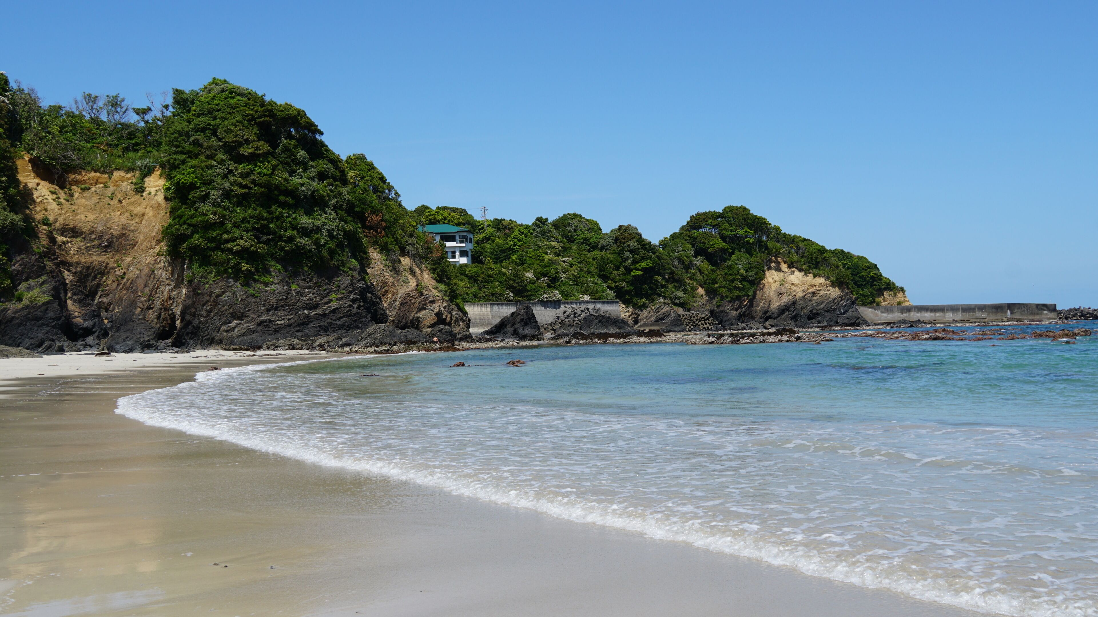 A beach with clear turquoise water, sandy shore, and a rocky cliff with dense greenery.