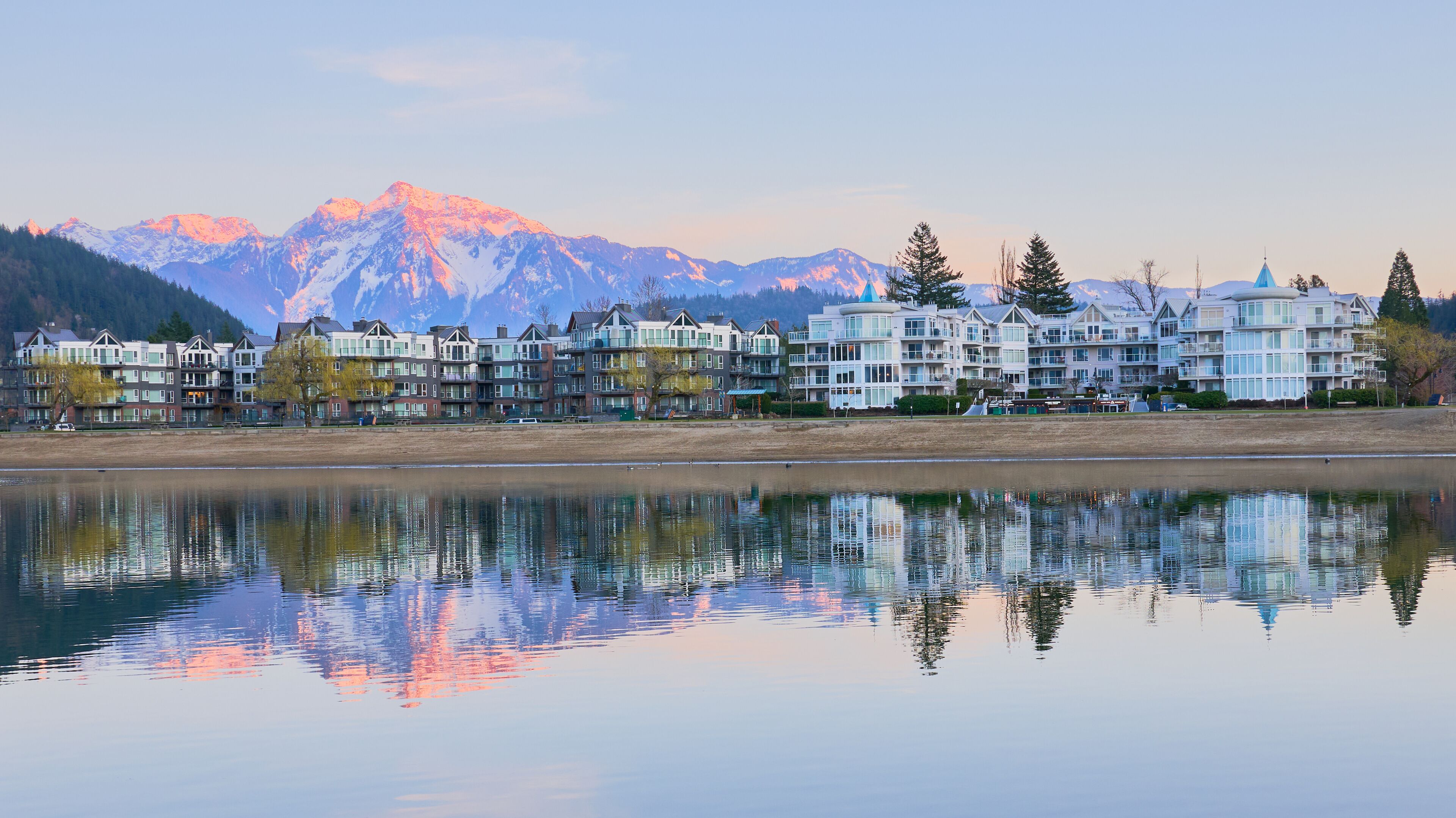 A lakeside view with a row of multi-story buildings, a calm water surface reflecting the structures, and snow-capped mountains in the background.