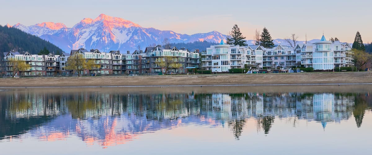 A lakeside view with a row of multi-story buildings, a calm water surface reflecting the structures, and snow-capped mountains in the background.