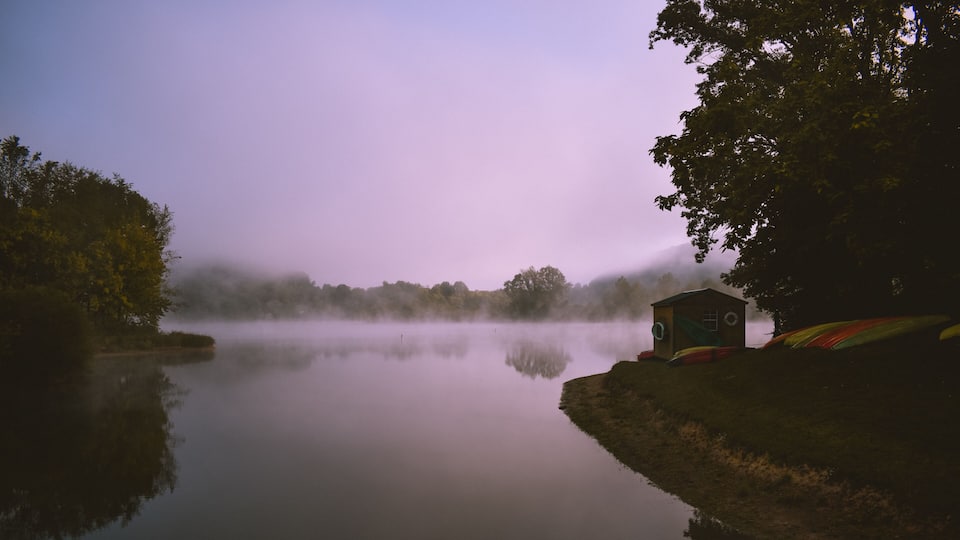 A calm lake with a wooden dock, a small cabin, and dense trees in the background.
