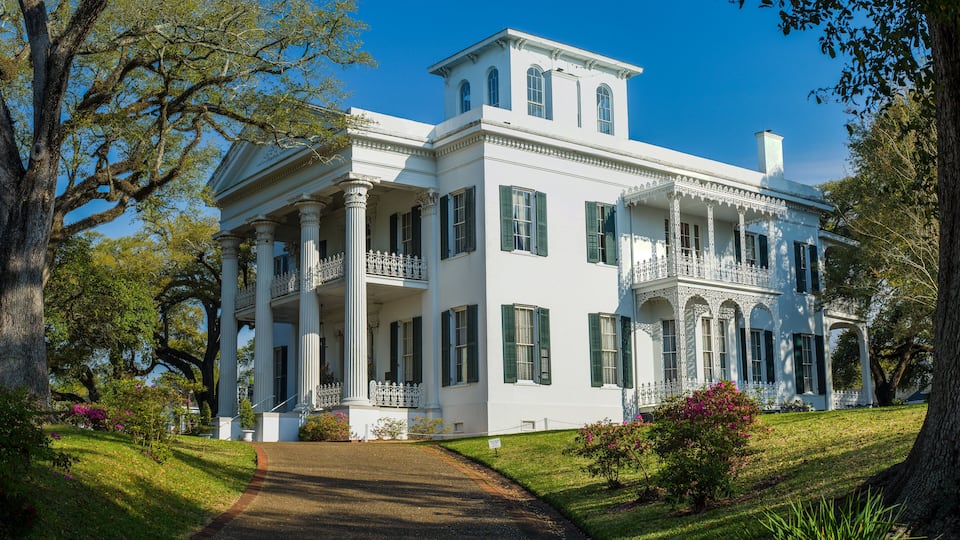 A large white mansion with green shutters, surrounded by lush greenery and trees.