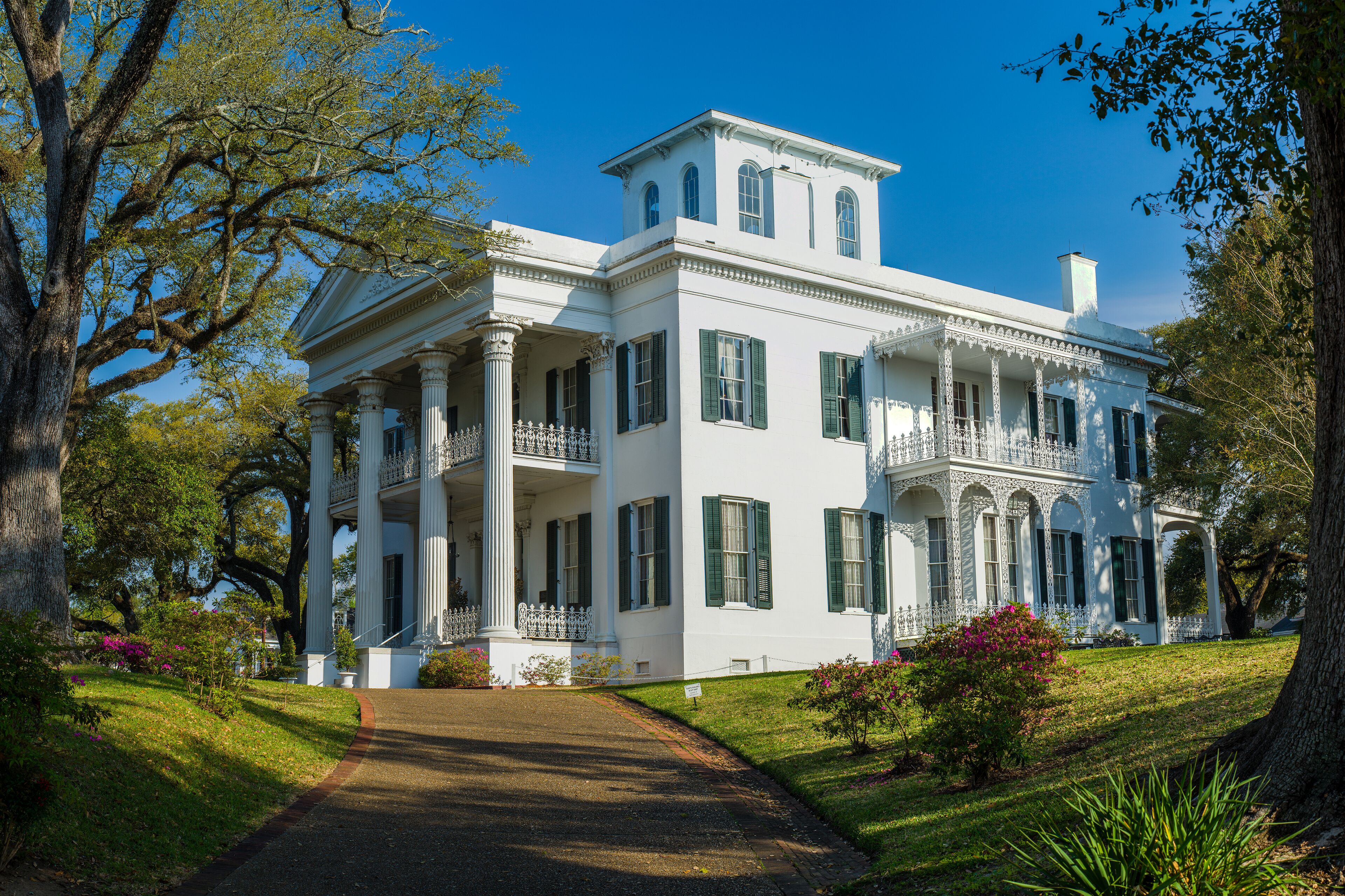 A large white mansion with green shutters, surrounded by lush greenery and trees.