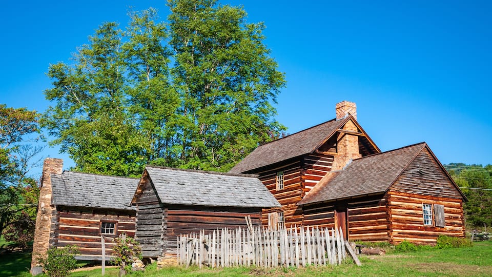 A rustic log cabin with a wooden fence, surrounded by lush green grass and trees.