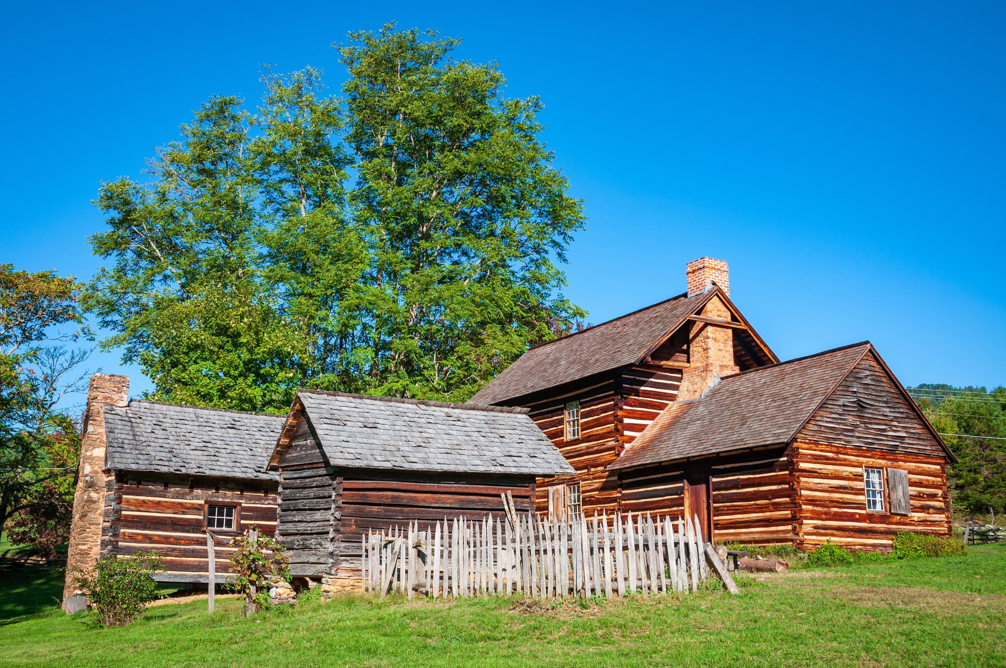 A rustic log cabin with a wooden fence, surrounded by lush green grass and trees.