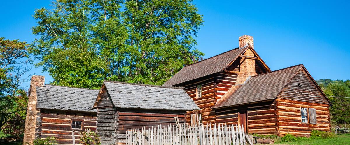 A rustic log cabin with a wooden fence, surrounded by lush green grass and trees.