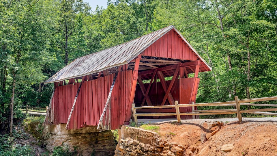A red covered bridge over a small stream, surrounded by dense green forest.