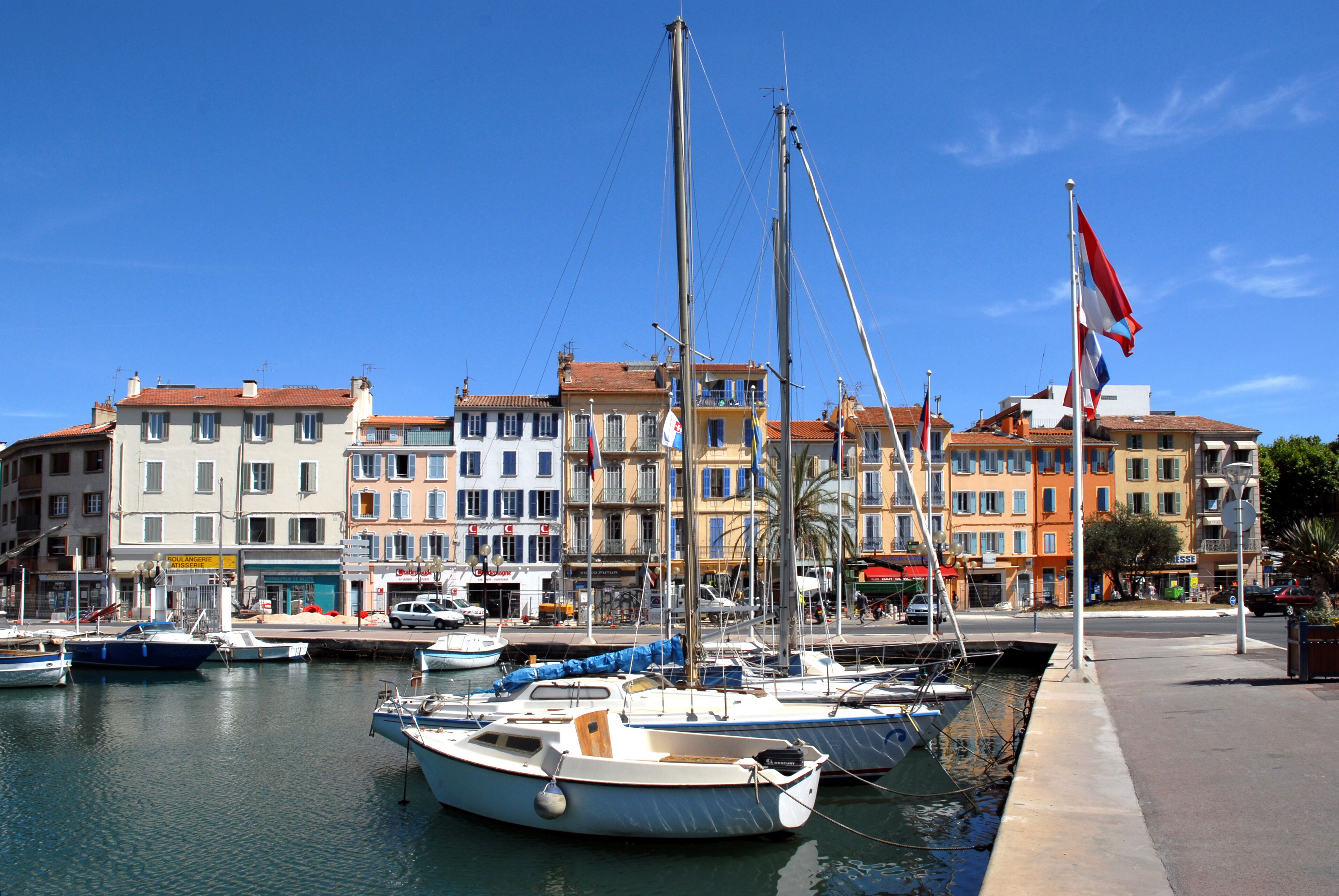 A marina with several boats docked, a flagpole with a flag, and colorful buildings in the background.