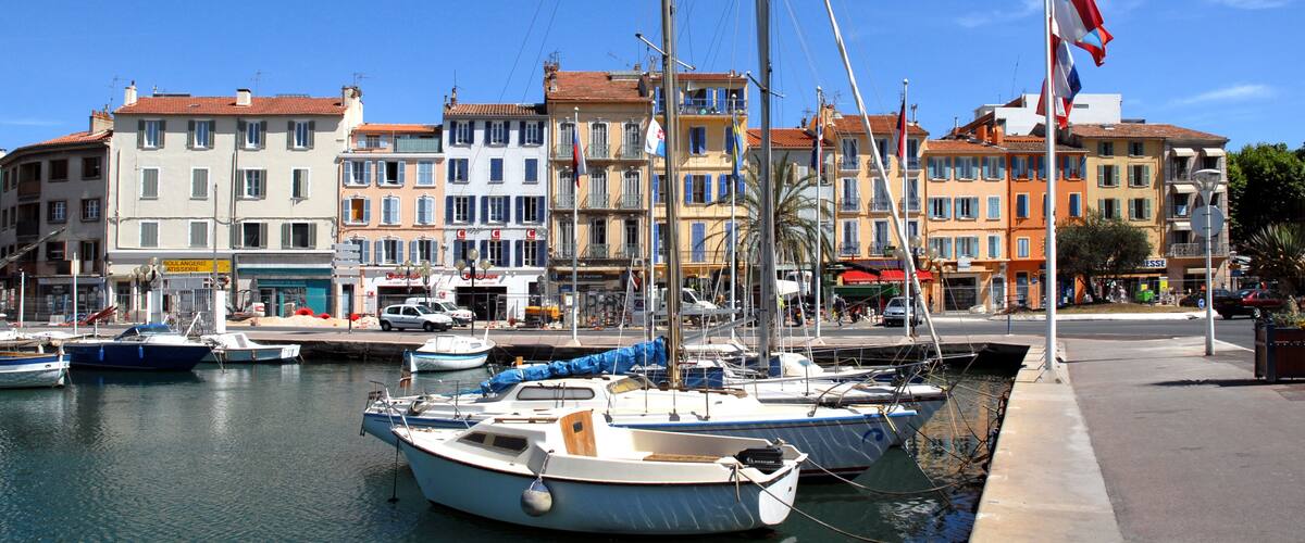 A marina with several boats docked, a flagpole with a flag, and colorful buildings in the background.