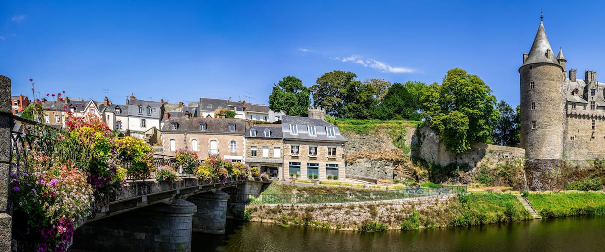A historic castle with a moat, a bridge with flower boxes, and a river.