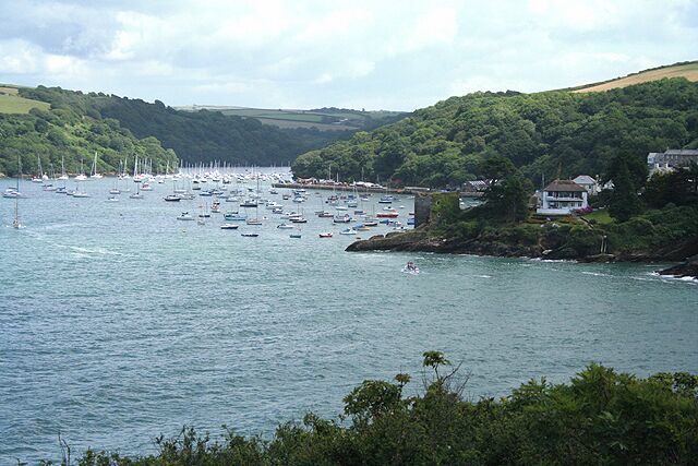 A coastal bay with numerous sailboats and yachts, surrounded by lush green hills and a few buildings on the shore.