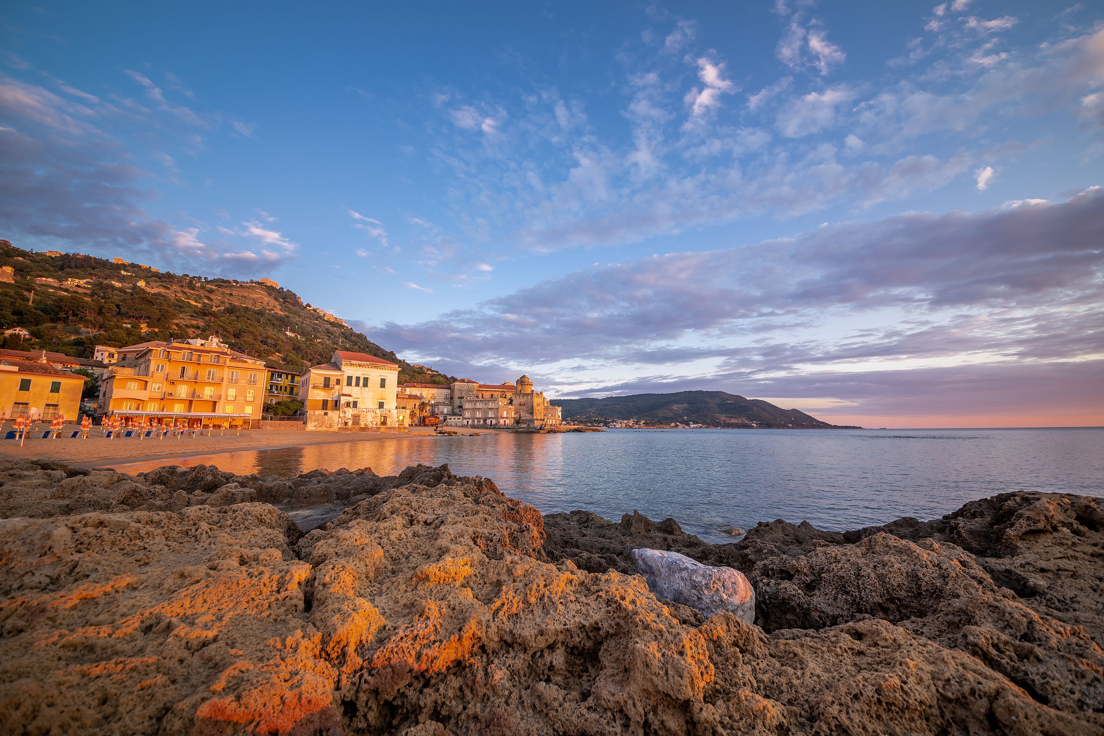 A coastal town with buildings on a hillside, rocky shoreline, and calm sea.