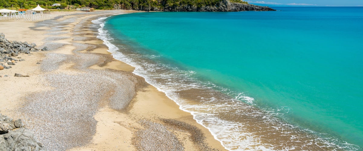 A beach with clear turquoise waters, a pebbly shore, and a forested hill in the background.