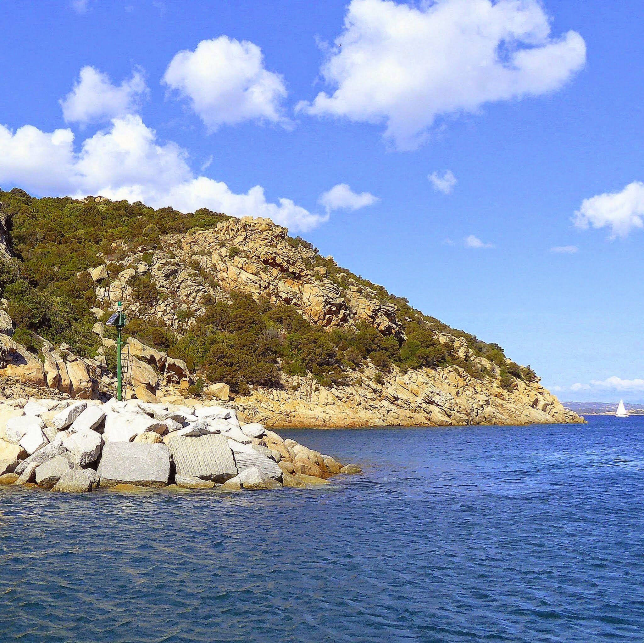 A rocky coastline with a lighthouse, clear blue sea, and a sailboat in the distance.
