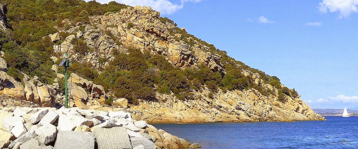 A rocky coastline with a lighthouse, clear blue sea, and a sailboat in the distance.